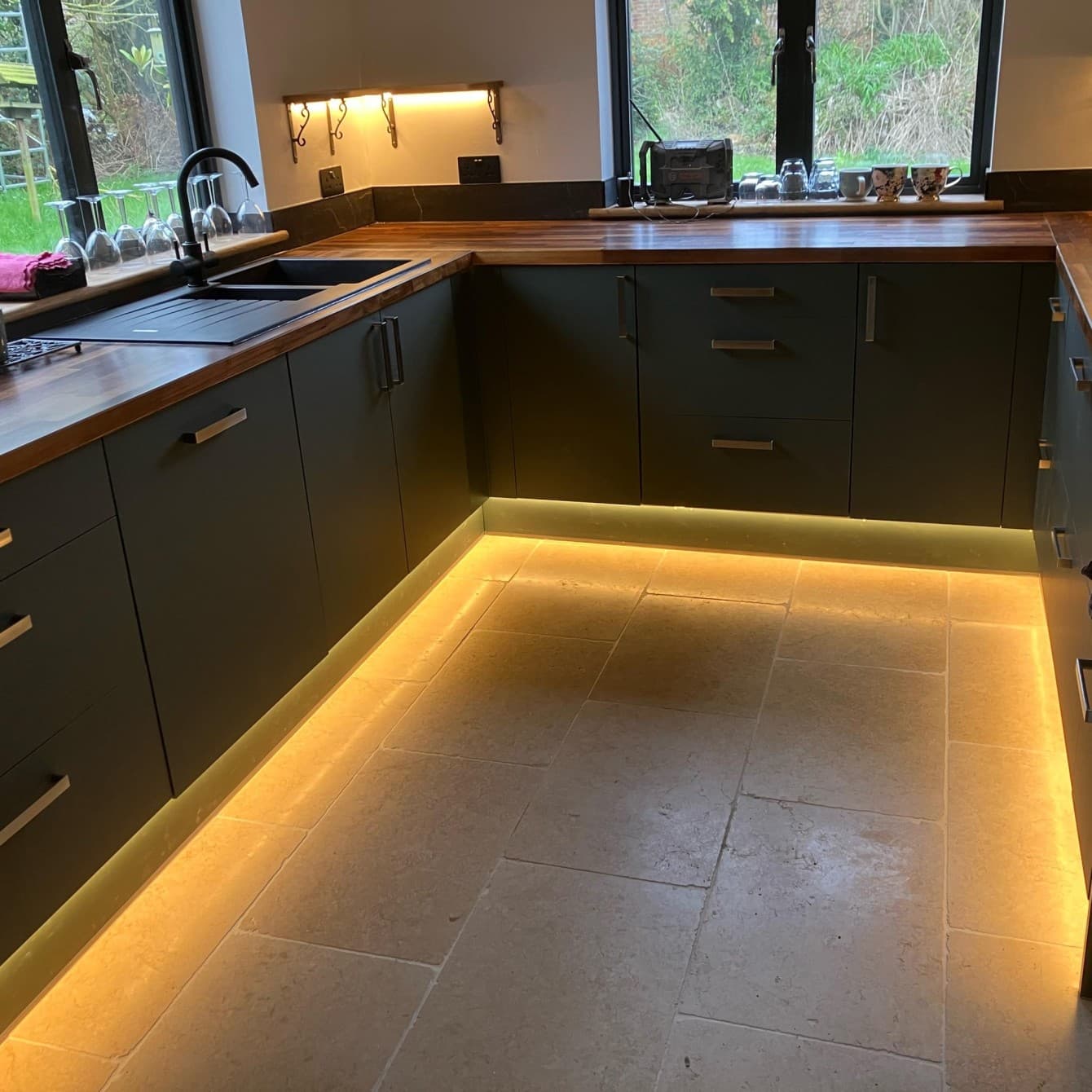 Warm plinth and under-cabinet LED lighting fitted in a dark green kitchen with wood worktops